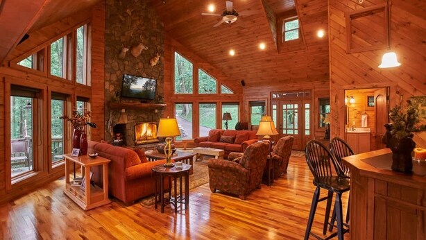 A spacious living room with a stone fireplace, wooden ceiling, and large windows offering views of the outdoors.