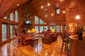 A spacious living room with a stone fireplace, wooden ceiling, and large windows offering views of the outdoors.