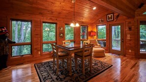 A wooden cabin interior with large windows, a dining table, and a sofa.