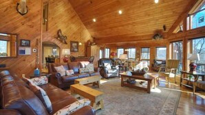 A spacious living room with a high ceiling, wooden walls, and large windows. There are leather sofas, a coffee table, and a fireplace.