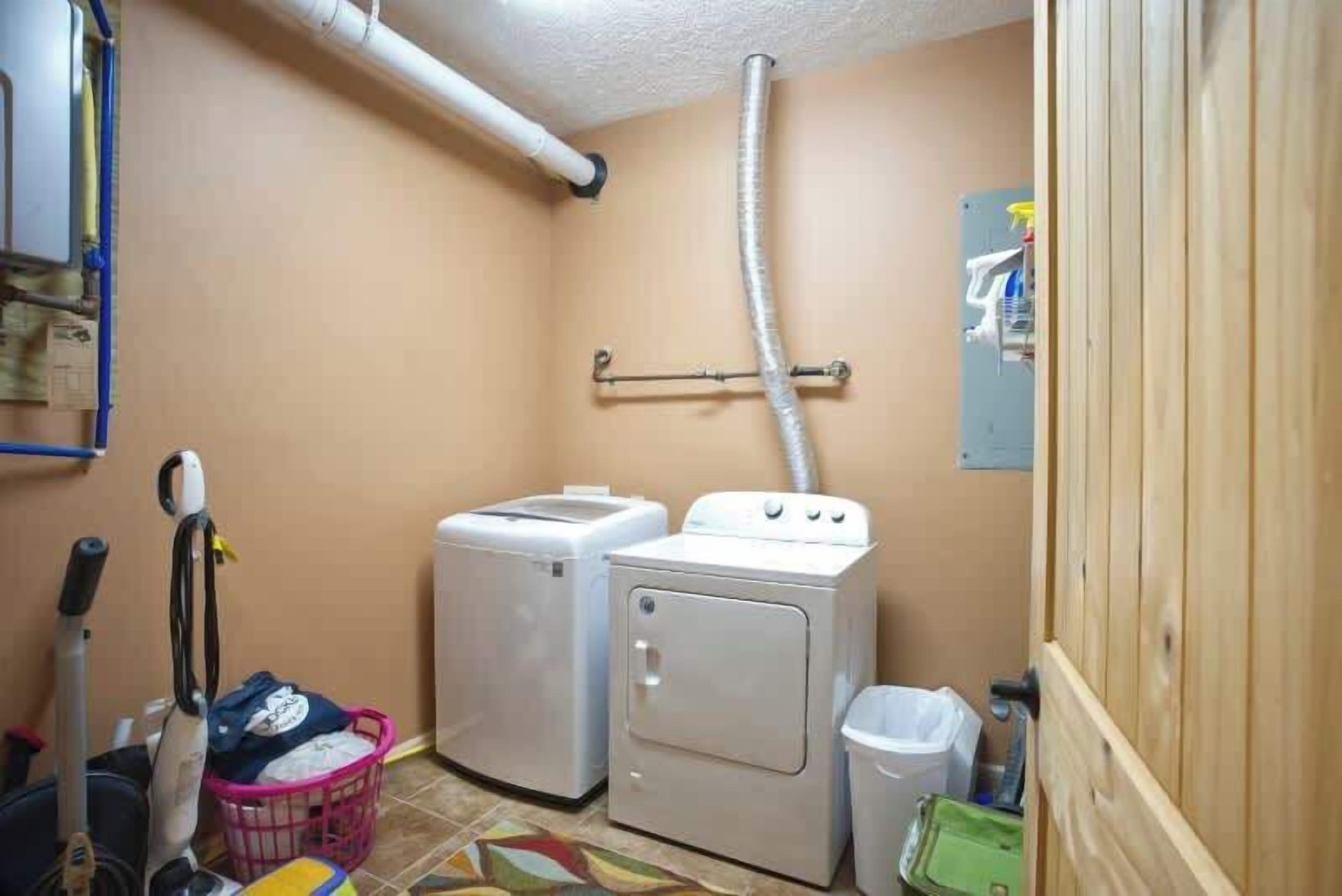 A laundry room with two washing machines, a dryer, a mop, a mop bucket, and a trash bin.