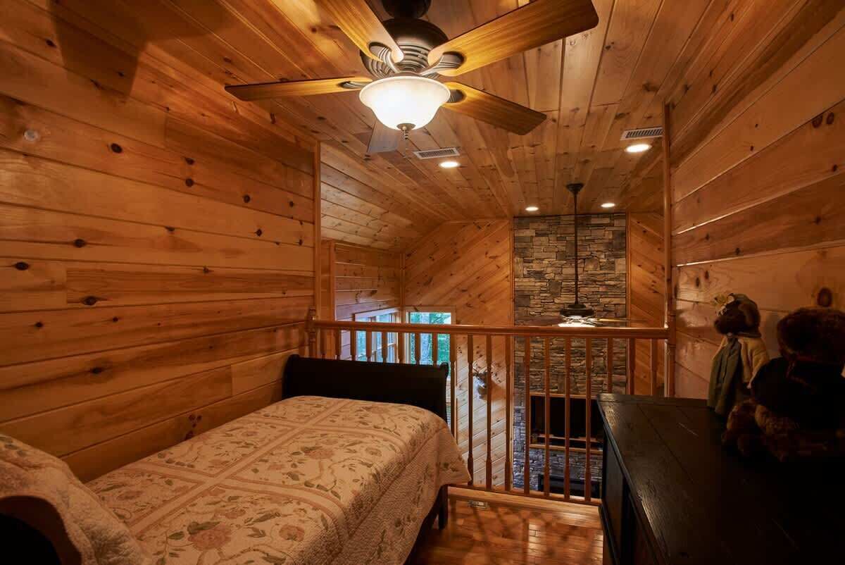 A cozy cabin interior with a wooden ceiling, a bed with a patterned bedspread, a stone fireplace, and a small seating area with a teddy bear.