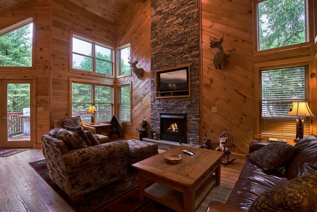 A cozy cabin interior with a stone fireplace, wooden walls, and large windows offering a view of the surrounding forest.