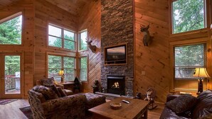 Ein gemütliches Blockhaus-Interieur mit Kamin aus Naturstein, Holzwänden und großen Fenstern mit Blick auf den umliegenden Wald.