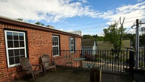 Une maison en briques avec une terrasse en bois, deux fauteuils Adirondack et une petite table.