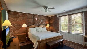 Une chambre avec un mur en briques, un lit en bois, deux tables de chevet avec des lampes, un ventilateur de plafond et une fenêtre avec des stores.
