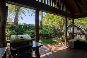 A covered outdoor area with a grill, wooden deck, and a view of a garden and house. - Bark Bungalow (Blowing Rock)