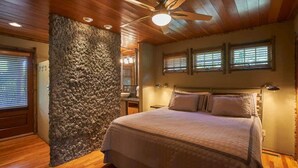 A bedroom with a large bed, wooden ceiling, stone wall, and two windows with blinds.