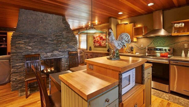 A modern kitchen with a stone fireplace, wooden ceiling, and stainless steel appliances.