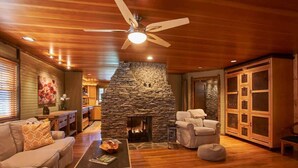 A cozy living room with a stone fireplace, wooden ceiling, and a ceiling fan.