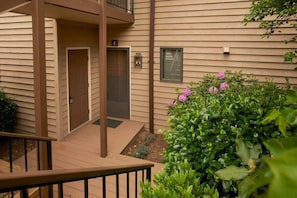 A residential building with a wooden deck, a door, and a small garden with pink flowers.