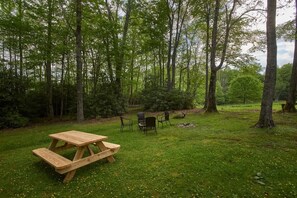 Une table de pique-nique avec des bancs, située dans une zone boisée, avec des arbres et un sol recouvert d’herbe.