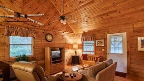 Ein gemütliches Blockhaus-Interieur mit Holzwänden, Deckenventilator, Kamin, Fernseher und einer Couch.