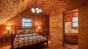 Ein gemütliches Schlafzimmer in einer Holzhütte mit einem Metallbettgestell, einem Fenster mit Vorhängen, einer Holzkommode und einem Badezimmer mit Waschbecken und Spiegel.