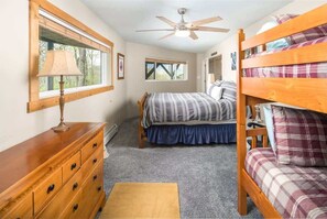 Una habitación compacta con literas, una cama individual, un aparador de madera, un ventilador de techo y una ventana con persianas.