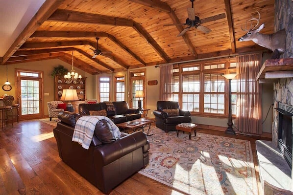 A spacious living room with wooden beams, leather sofas, a fireplace, and large windows.