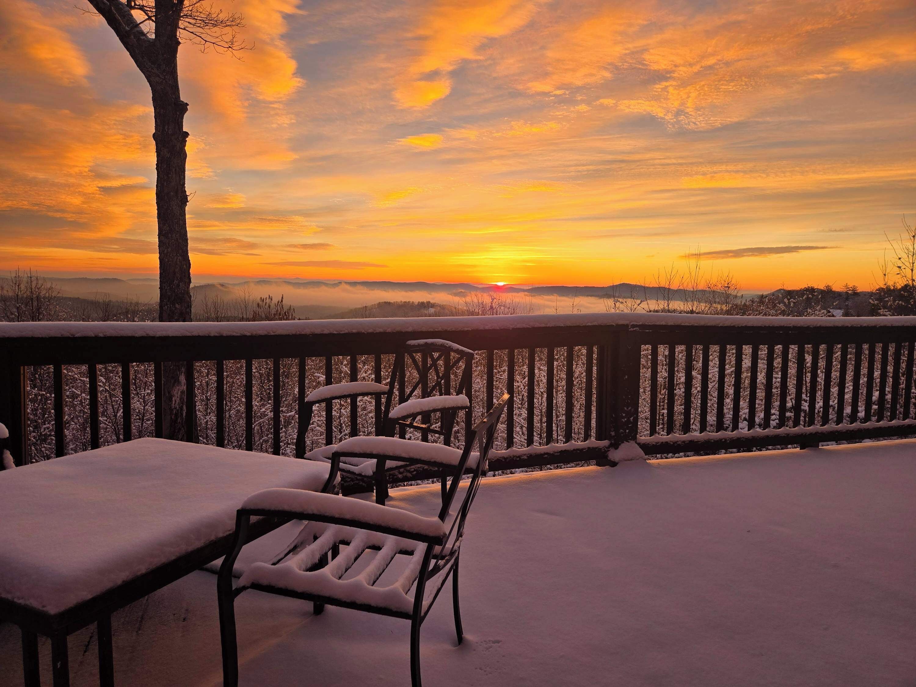 Ein mit Schnee bedeckter Balkon mit Tisch und Stühlen, mit Blick auf einen Sonnenuntergang mit Bergen im Hintergrund.
