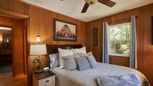Ein Schlafzimmer mit Holzwandverkleidung, einem Bett mit vielen Kissen, einem Nachttisch mit Lampe und einem Deckenventilator.
