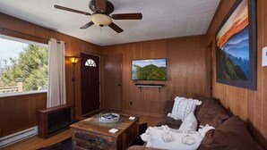 Ein gemütliches Zimmer mit Holzwänden, einem Deckenventilator, einem Kamin, einem braunen Sofa und einem großen Fenster mit Blick auf Bäume.
