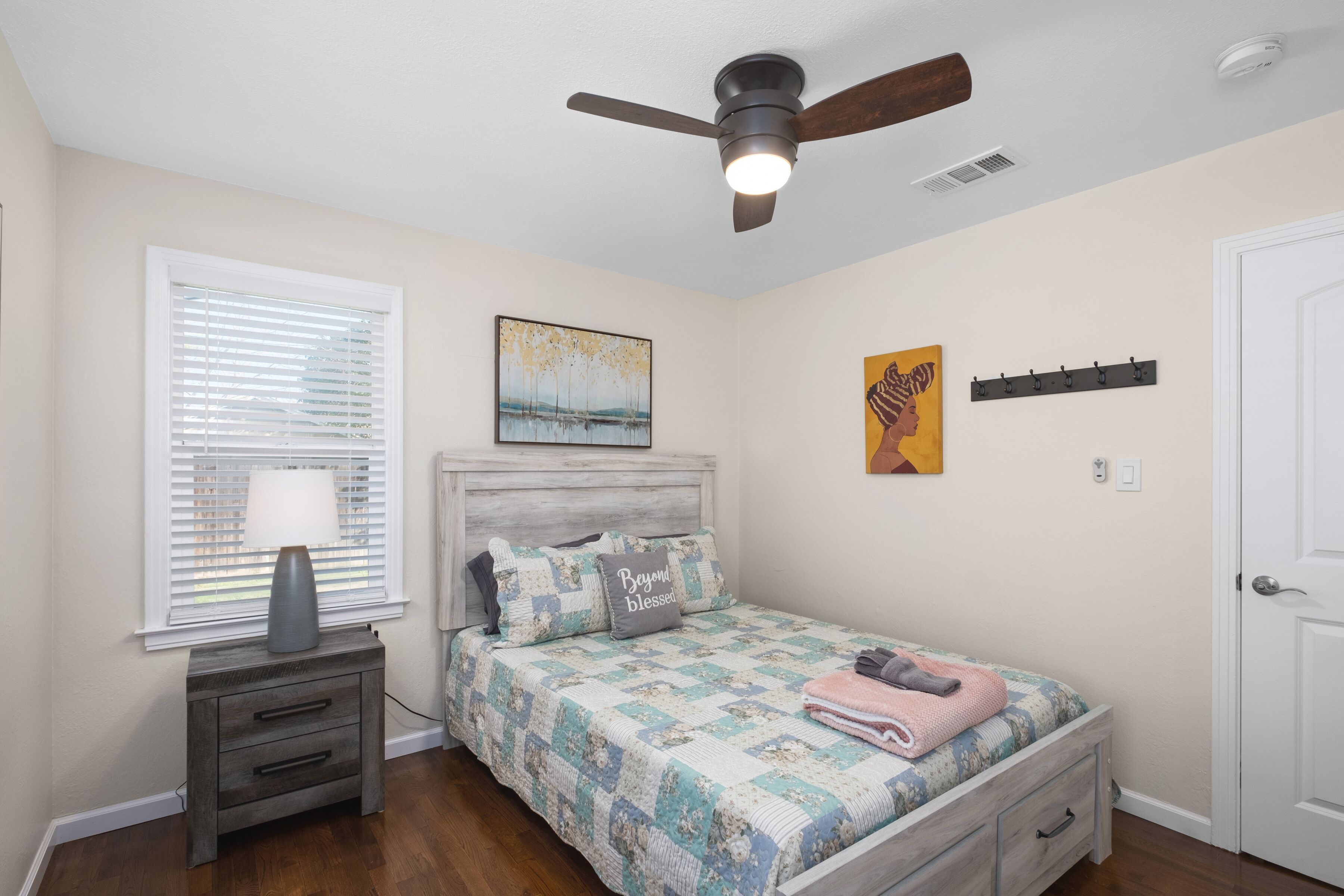 A bedroom with a bed, a nightstand, a ceiling fan, a window with blinds, and a door.