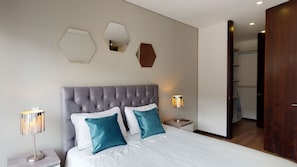 A bedroom with a tufted headboard, a large bed with white bedding and teal pillows, two bedside lamps, and a wooden door leading to a hallway. - APARTMENT LUXOR 8FLOOR-AREA97-803 (Bogotá)