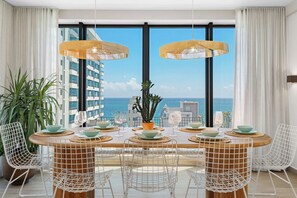 A dining area with a table set for four, surrounded by white wire chairs, with a view of the ocean through large windows. - HiBird- Apartment and Suites Hotel (San Juan)