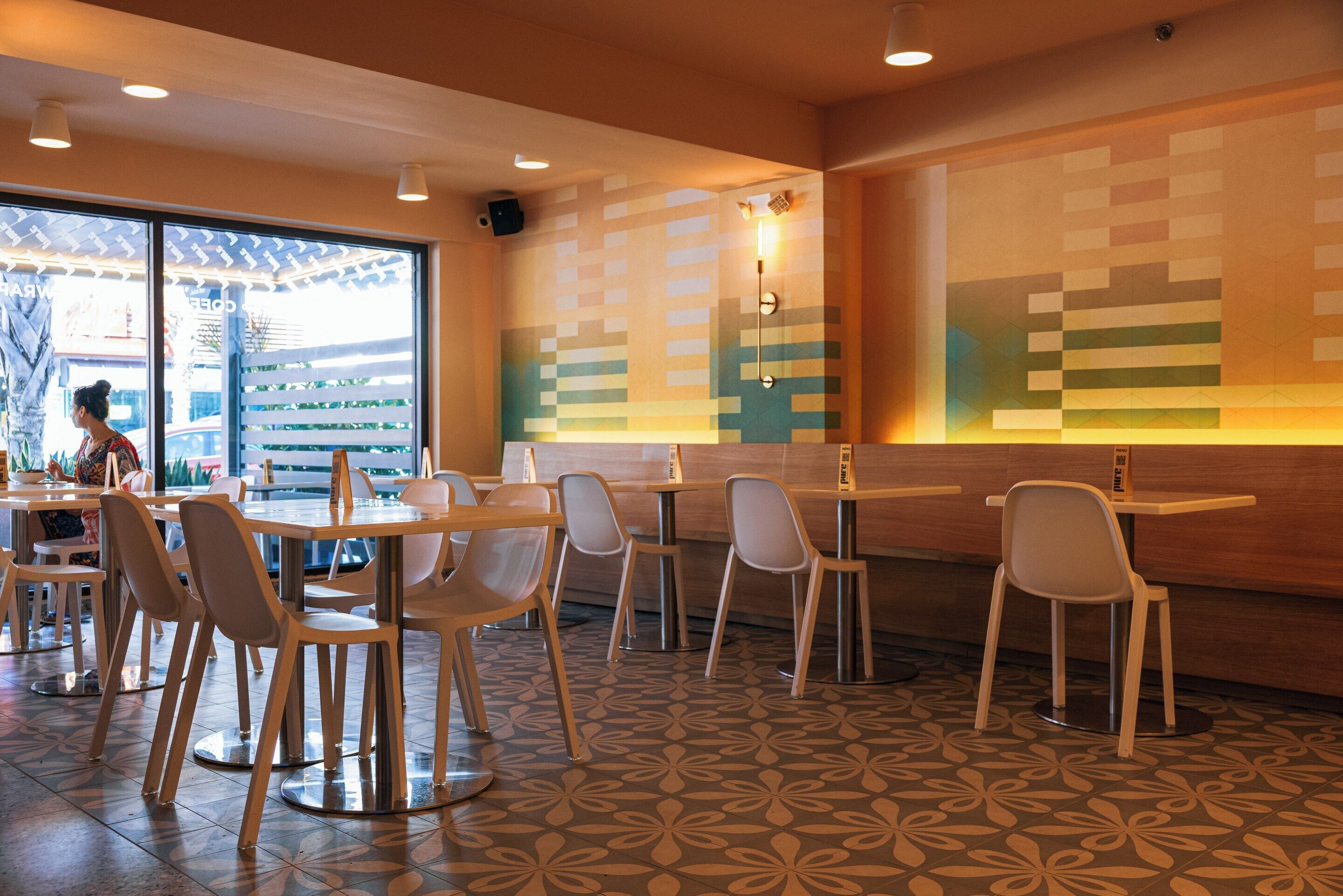 Un intérieur de restaurant moderne, avec des tables et des chaises rondes, un sol à motifs et une grande fenêtre donnant sur l’extérieur.