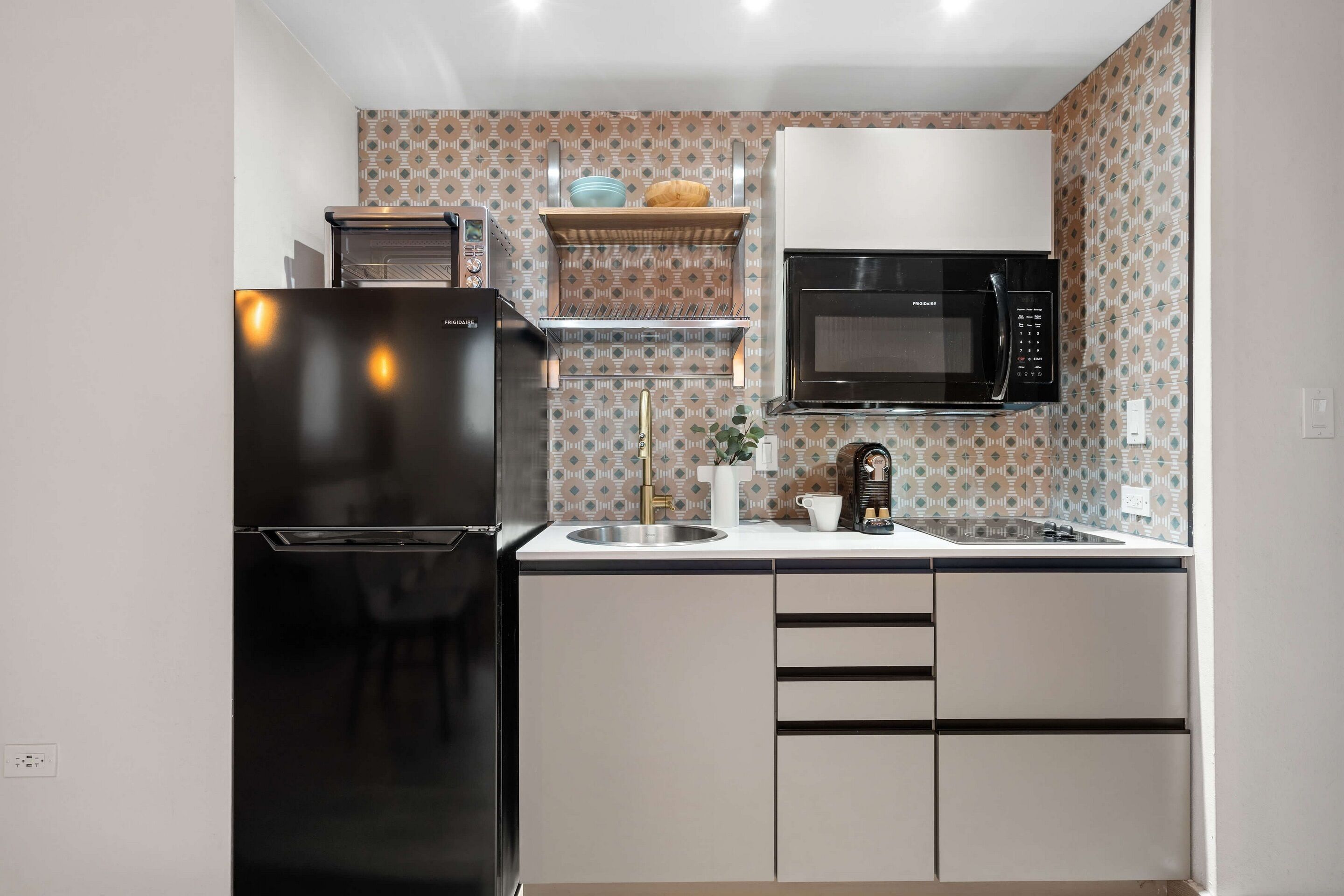 A compact kitchen with a black refrigerator, a microwave, and a sink. There is a shelf above the sink with a croissant and a blue bowl.