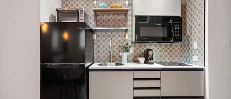 Una cocina compacta con un refrigerador negro, un microondas y un lavamanos. Hay una repisa sobre el lavamanos con un croissant y un tazón azul.