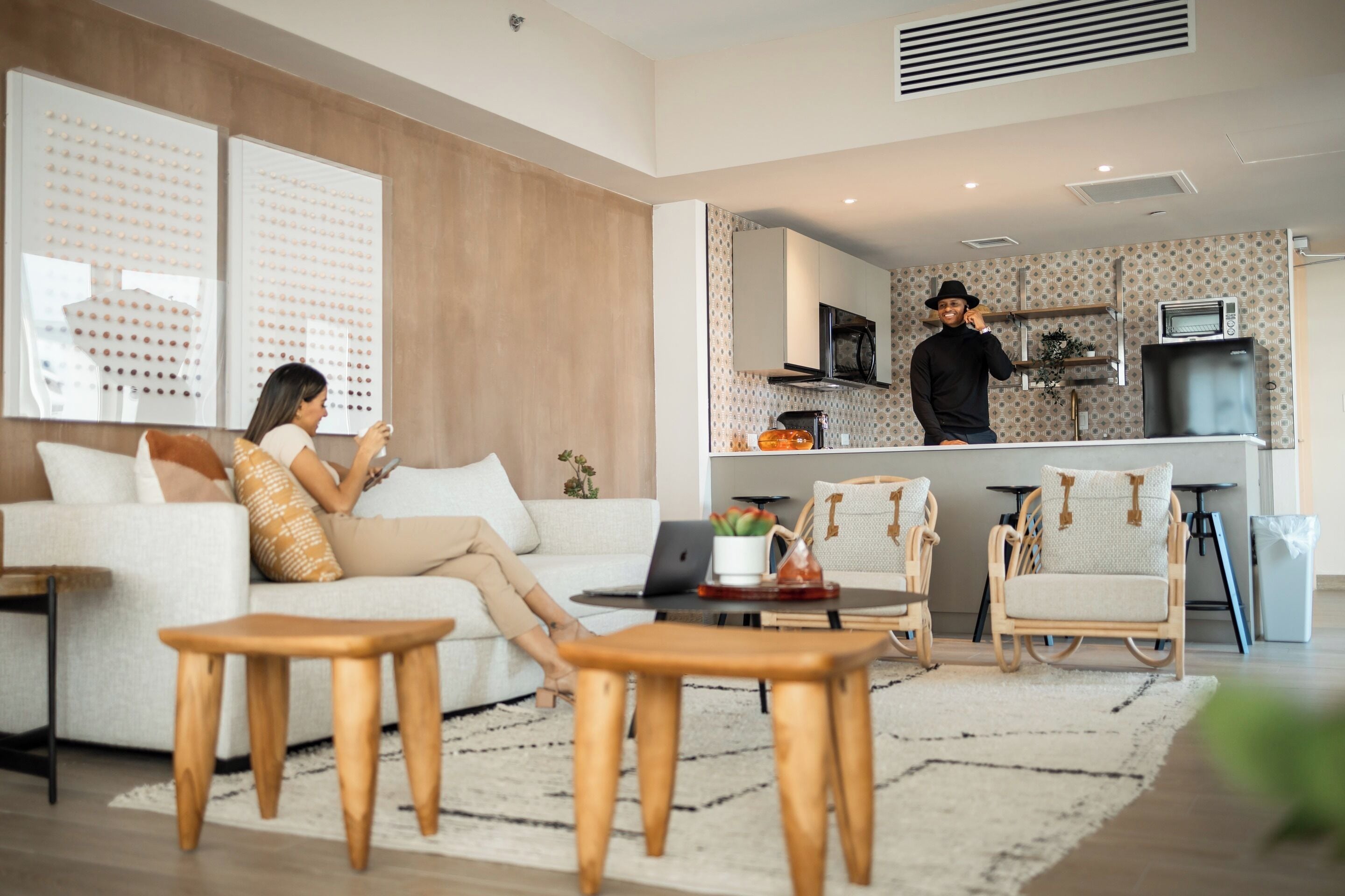 Un salon moderne avec un canapé, une table basse et des fauteuils. On aperçoit, au fond, un coin cuisine avec un four à micro-ondes et un réfrigérateur.
