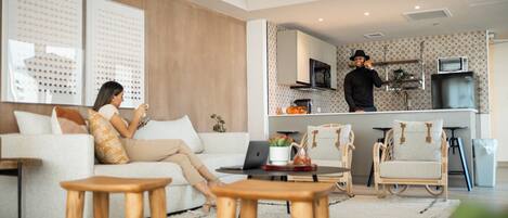 A modern living room with a sofa, coffee table, and chairs. A kitchen area is visible in the background with a microwave and refrigerator.