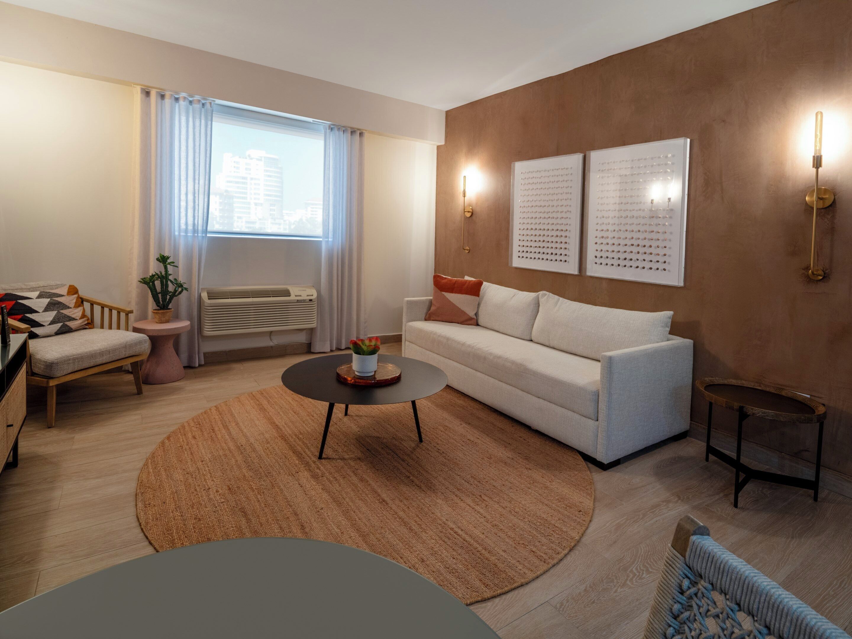A modern living room with a sofa, coffee table, and a small side table. There is a window with curtains, a wall-mounted air conditioner, and two wall sconces.