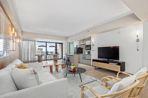 A modern living room with a sofa, coffee table, and wicker chairs. A TV is mounted on the wall, and a kitchen is visible in the background.