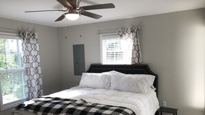 Une chambre à coucher avec un lit, un ventilateur de plafond, une fenêtre avec des rideaux et une petite table de chevet.