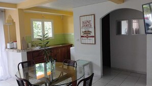 A dining area with a glass table, chairs, a small island with a plant, and a kitchen in the background.