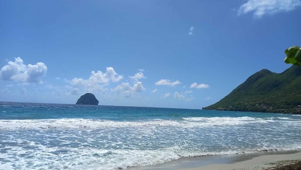 A beach with waves, a distant island, and a green hill.