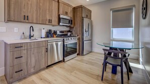 Una cocina moderna con armarios de madera, electrodomésticos de acero inoxidable, una mesa de comedor de vidrio y una ventana con persianas.