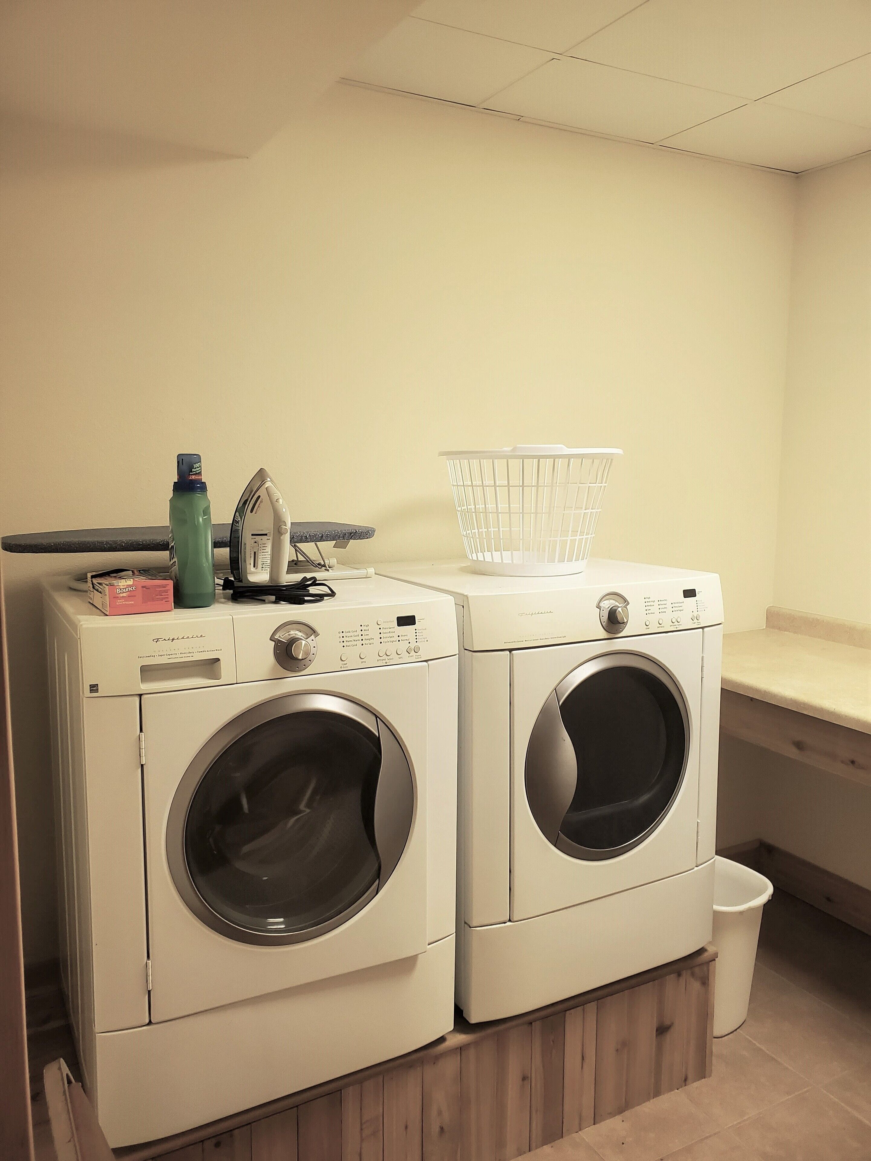 A laundry area with two front-loading washing machines, a drying rack, and a trash bin.