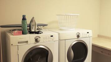 A laundry area with two front-loading washing machines, a drying rack, and a trash bin.