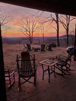 Ein Außenbereich mit Sitzgelegenheiten, Tischen und Blick auf eine Landschaft mit Bäumen und einem Sonnenuntergang.