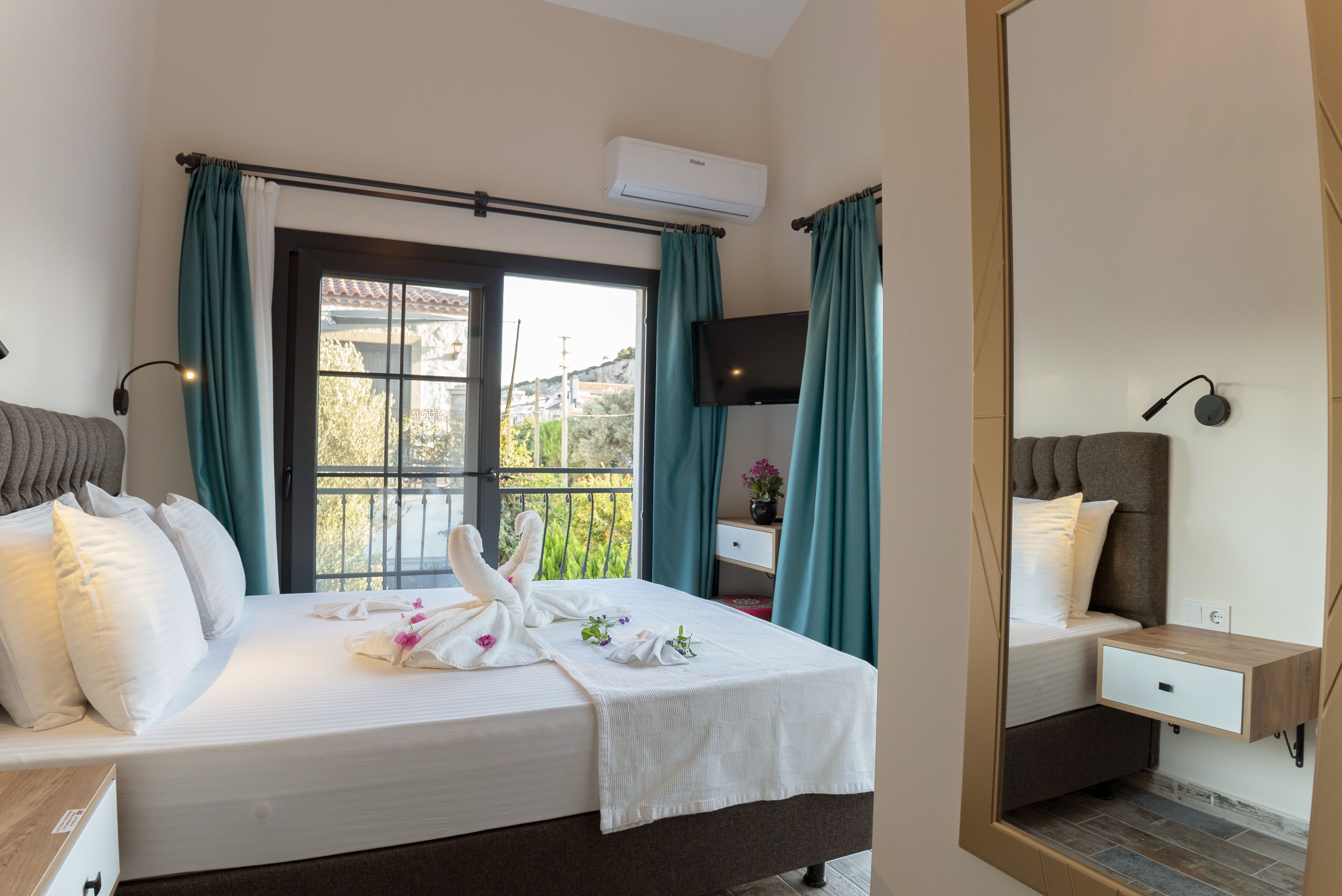 A hotel room with a bed, a bedside table, a nightstand, a mirror, a window with curtains, and an air conditioning unit.