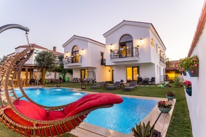 Un área junto a la piscina con una silla de mimbre, una piscina y un edificio de dos plantas con balcones.