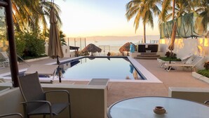 Un espace piscine aménagé avec des chaises longues et une table, offrant une vue sur une étendue d’eau calme où l’on aperçoit un voilier au loin.