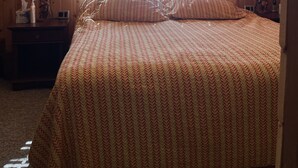 A cozy attic bedroom with a sloped ceiling, a bed with patterned bedding, a nightstand with a lamp, and a window with blinds. - Cozy cabin in Duck Creek (Duck Creek Village)