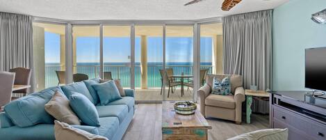 A spacious living room with a large sofa, a coffee table, and a TV. There is a balcony with a view of the ocean.