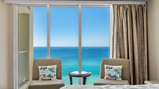 A hotel room with a large sliding glass door leading to a balcony with a view of the ocean. There are two armchairs with pillows, a small round table, and a bed with a white bedspread. - Long Beach Resort Tower 1 Unit 1303 (Panama City Beach)