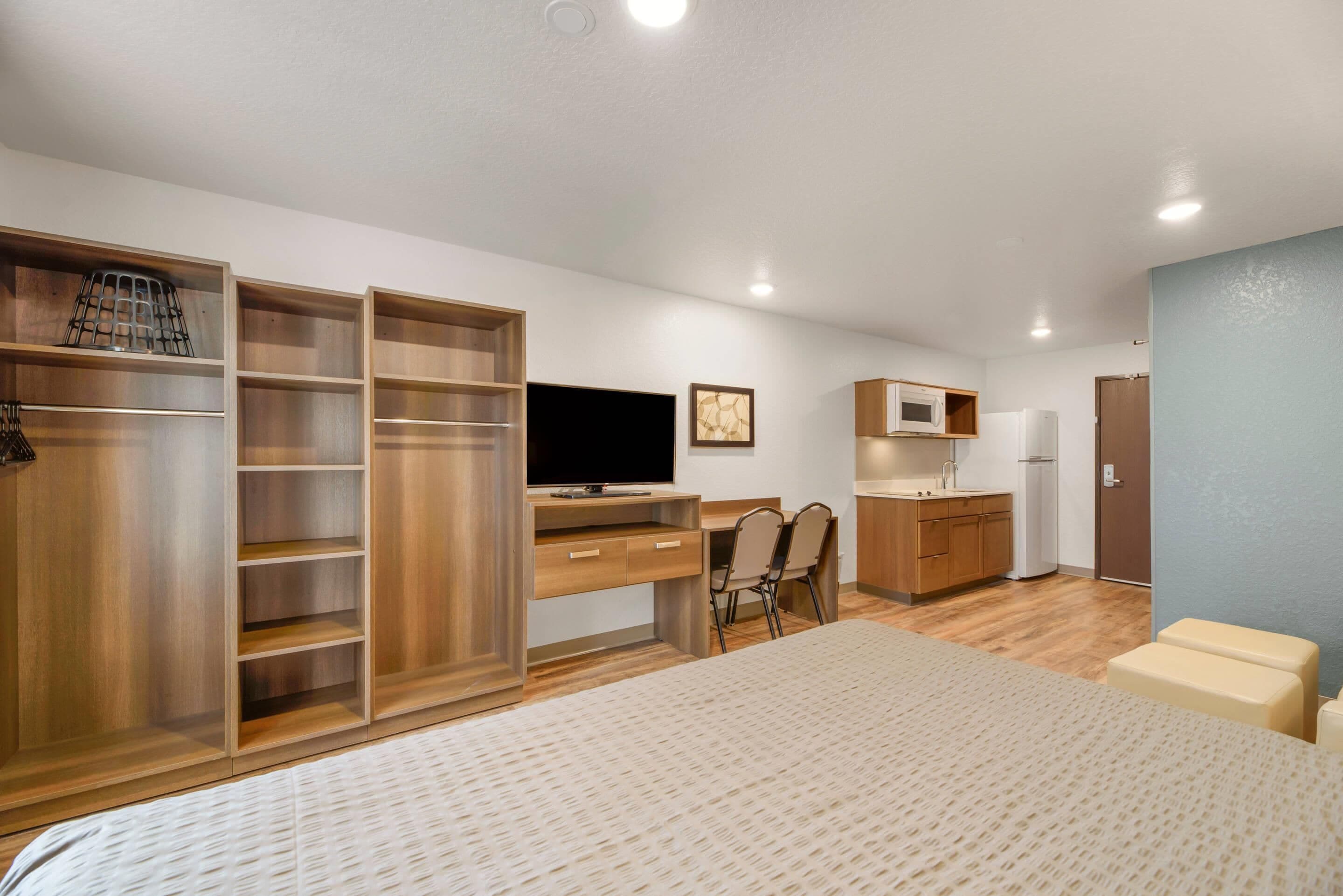 A compact room with a bed, a desk with a computer, a television, a small kitchenette, and a wardrobe.