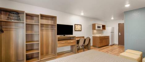 A compact room with a bed, a desk with a computer, a television, a small kitchenette, and a wardrobe.