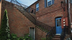 Un bâtiment en briques de deux étages, avec une porte bleue, un escalier doté de rampes noires et un petit jardin agrémenté d’arbustes et de fleurs.