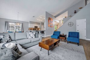 A spacious living area with a gray sofa, blue armchairs, and a wooden coffee table. A kitchen is visible in the background, and a staircase leads to another level.